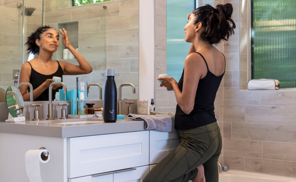 Woman applying face lotion in the bathroom mirror with Water.io bottle sitting on counter.