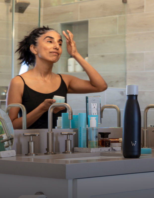 Woman applying face cream in bathroom mirror with Water.io bottle sitting on counter.